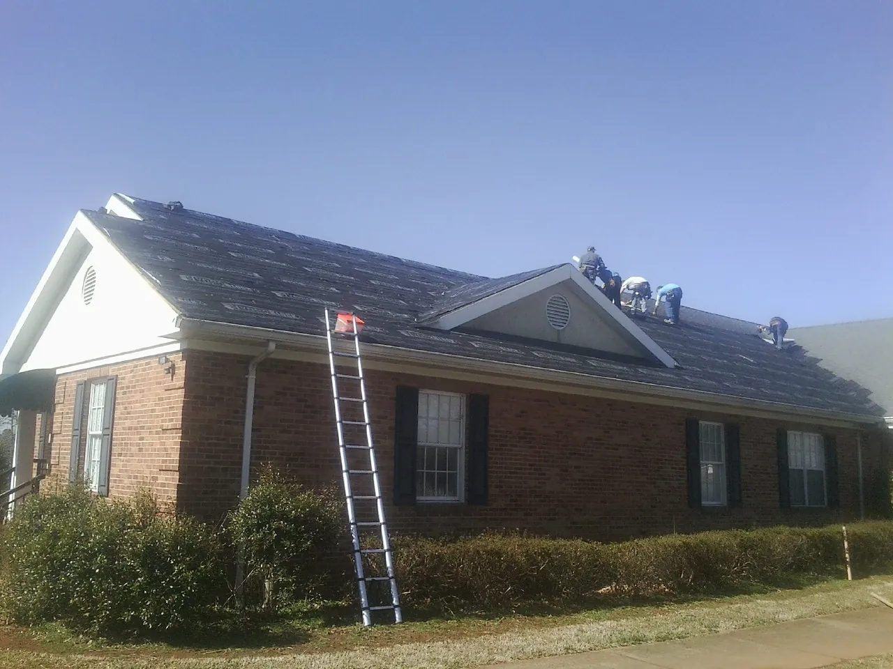 Crew tearing shingles off a brick ranch roof with a ladder set — Metal Roof Repair work by our crew in Moretown