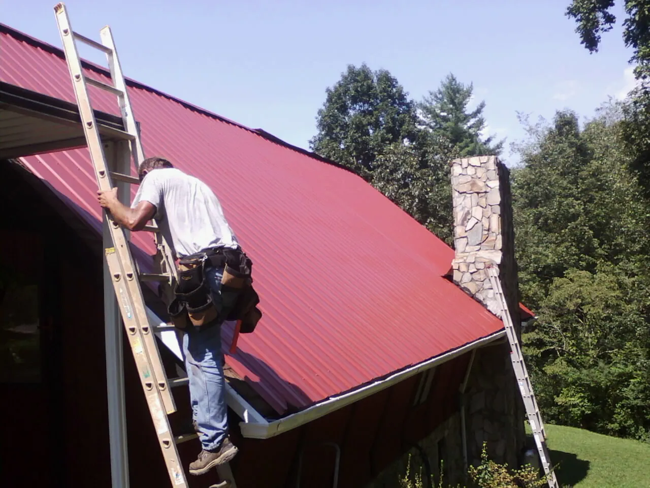 Roofer on a ladder beside a red metal roof and stone chimney — Soffit & Fascia Repair by Roofing Brothers in Moretown